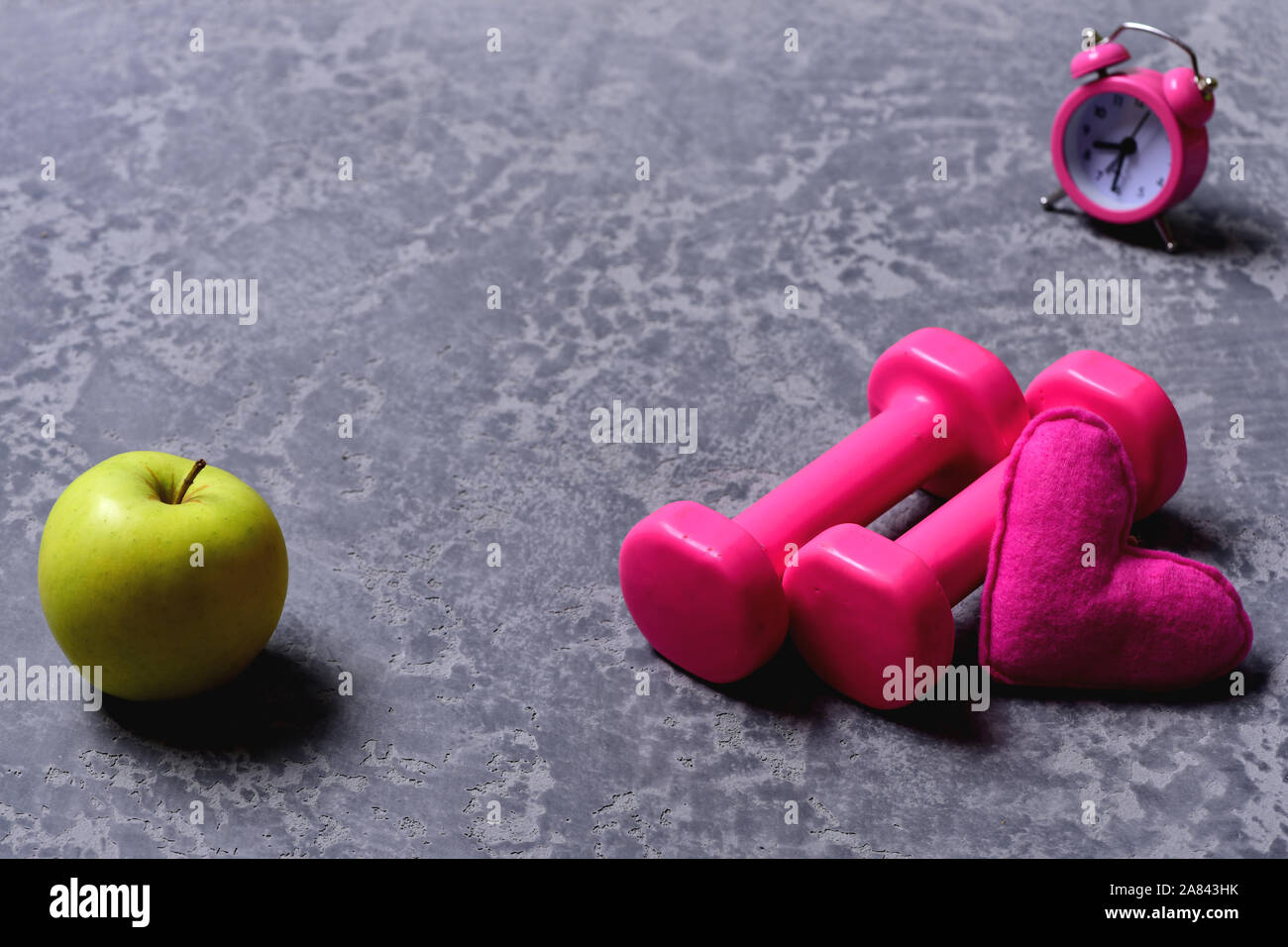 Weibliche Stil und Morgen Workout Konzept. Hanteln in rosa Farbe neben Apple, Wecker und Soft Toy Herz auf grauem Hintergrund. Herz-, Apple- und Kunststoff barbells. Begeisterung für den Sport und frühes Training Stockfoto