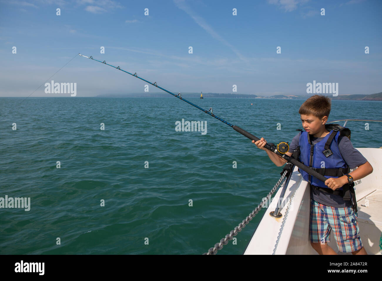 Ein kleiner Junge, der eine Angelrute auf einem Boot hält, während er an einem sonnigen Tag in Pembrokeshire, Wales angeln kann. Stockfoto