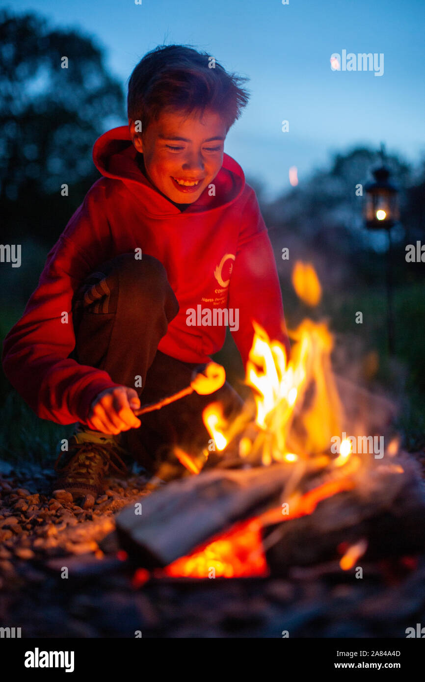 Ein kleiner Junge lächelt, während er beim Campen Marshmallows auf einem offenen Feuer kocht. Stockfoto