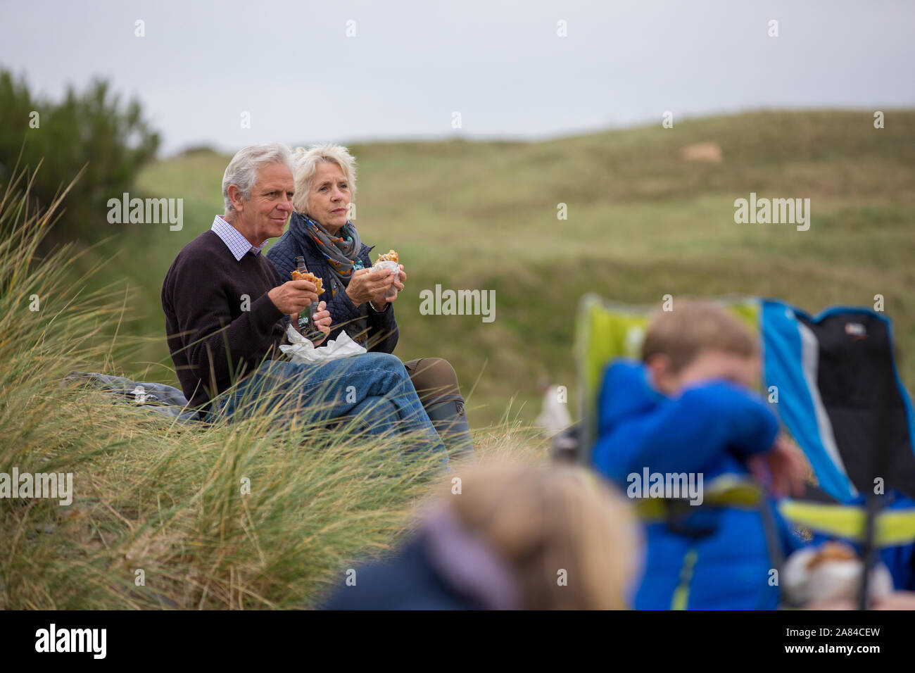 Großeltern essen eine Pastete in den Dünen sitzen und beobachten ihre Enkelkinder zu spielen. Stockfoto