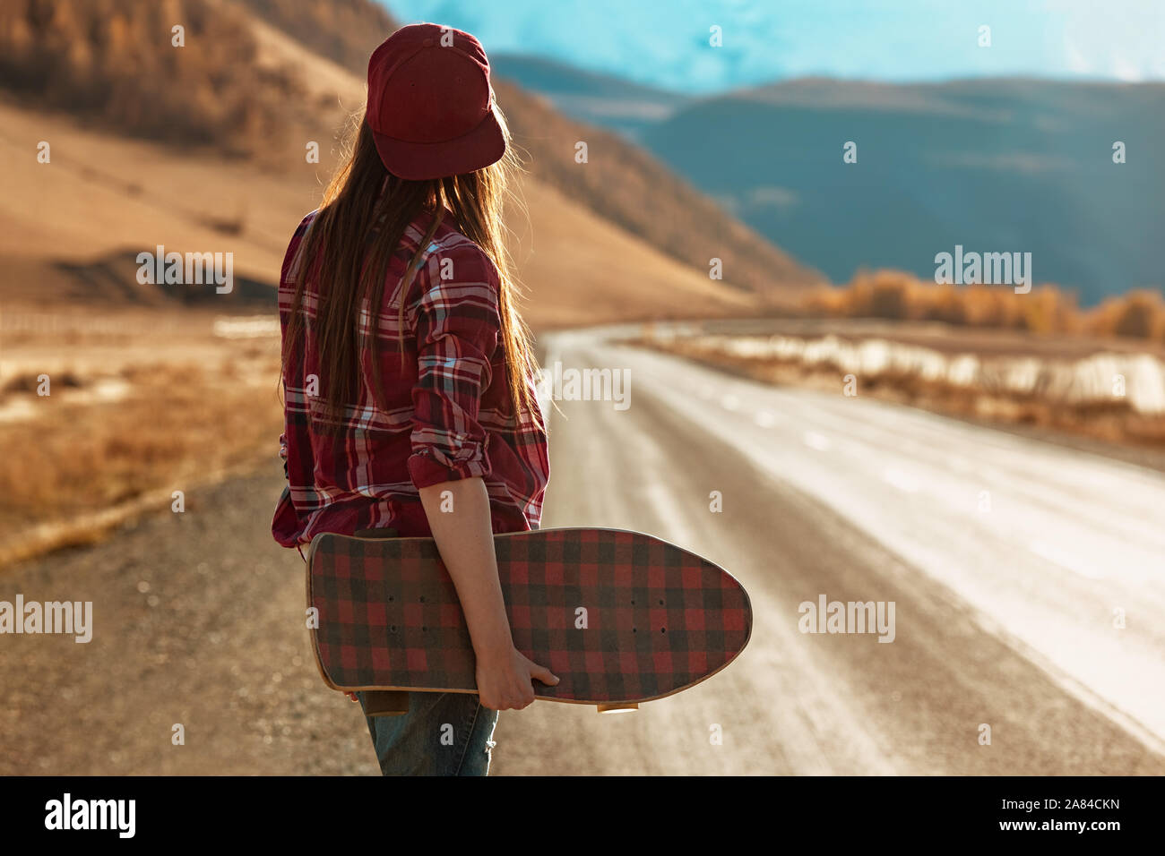 Hübsches Mädchen steht mit Longboard in Händen an der geraden Straße gegen Berge und den Sonnenuntergang Stockfoto