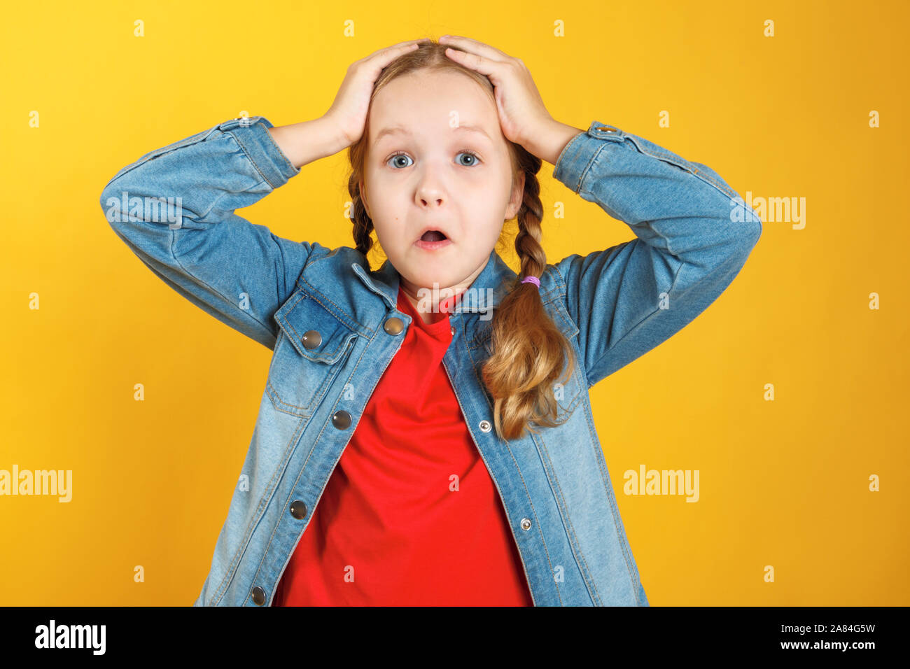 Lustiges kleines Mädchen hält die Hände hinter den Kopf auf einem gelben Hintergrund. Das Kind ist erstaunt, überrascht, emotional. Stockfoto