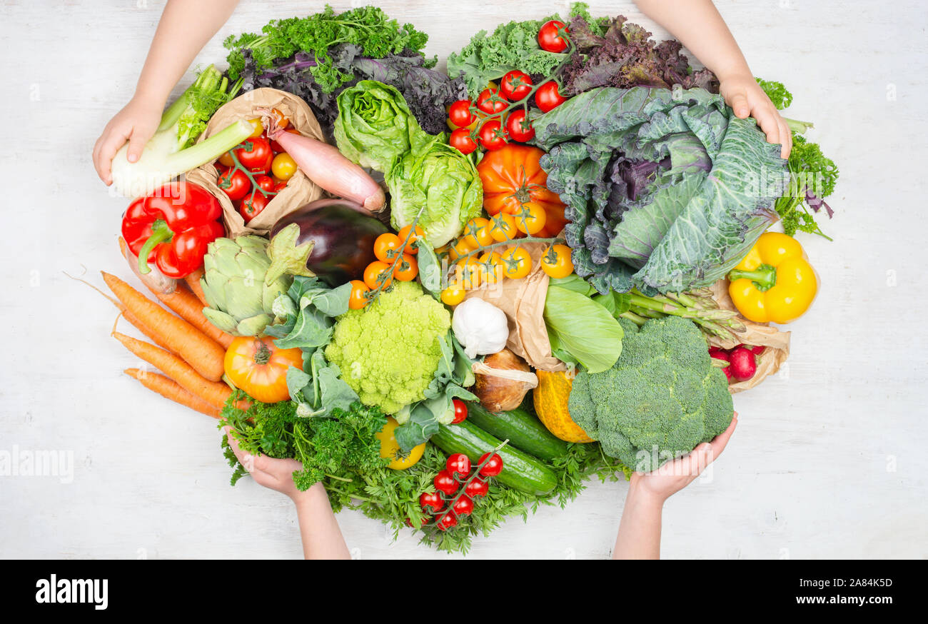 Das gesunde Essen. Child's Hände, die Rainbow Gemüse in einer Form des Herzens auf weissem Holztisch Tomaten Kohl Grüns Möhren Brokkoli, Ansicht von oben, auf weissem Holztisch, selektiver Fokus Stockfoto