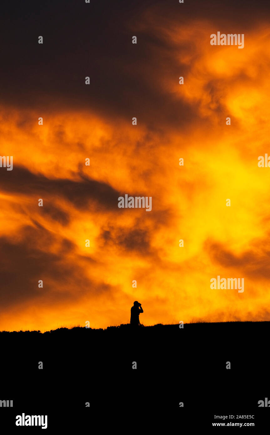 Ein Mann mit einem Fernglas gegen eine leuchtende Orange bewölkte Himmel bei Sonnenuntergang Stockfoto