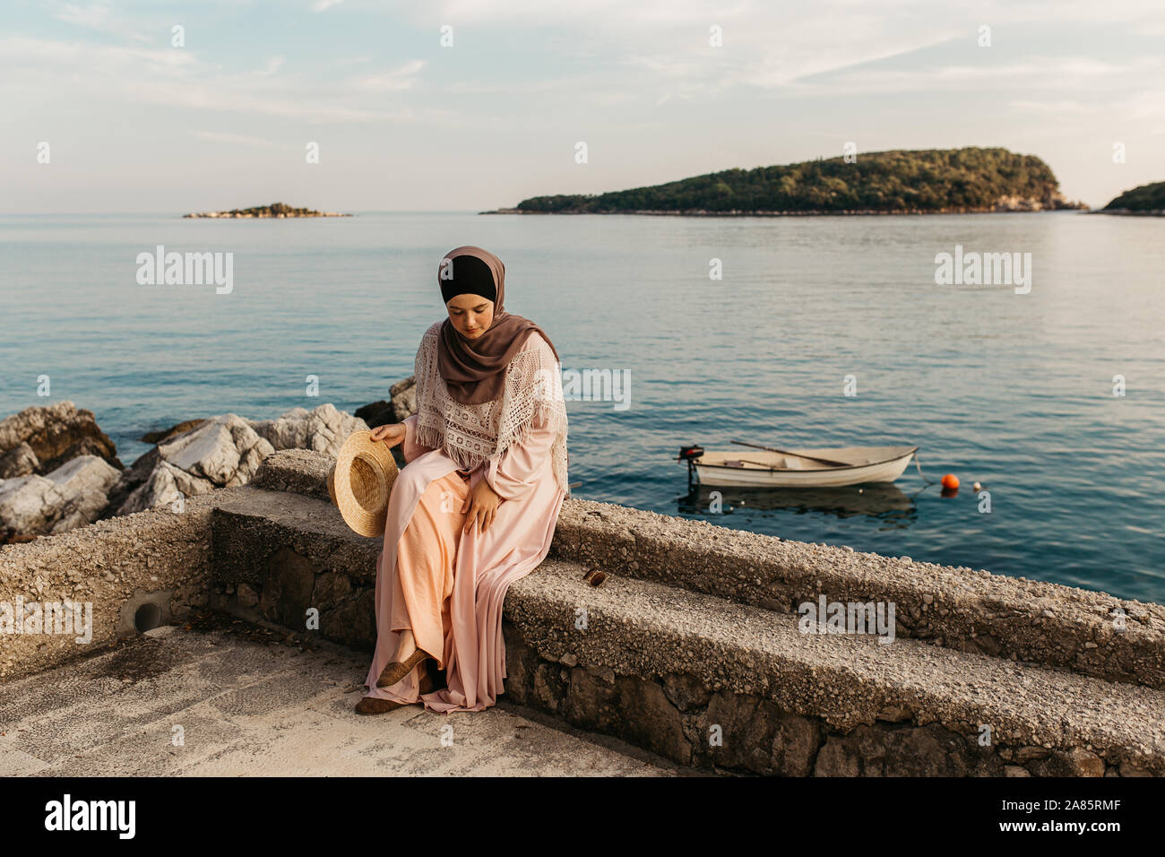 Porträt der jungen europäischen muslimischen Frauen mit Kopftuch sitzen am  Strand mit Hut in der Hand. Sie ist glücklich und entspannt. Das Meer ist  im Hintergrund Stockfotografie - Alamy