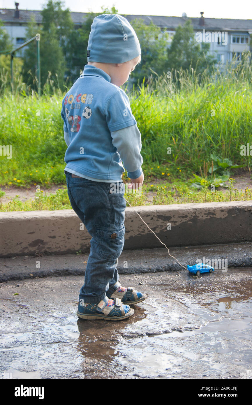 Ein 3-jähriger weißer Junge in einer blauen Hut, Jeans und Sandalen spielt an einem kühlen Sommertag mit einem überholten Fernbedienung Auto. Das Kind steht mit dem Rücken zum Betrachter auf einem Hintergrund von grünem Gras. Juli 2019 - Russland, Novodvinsk Stockfoto