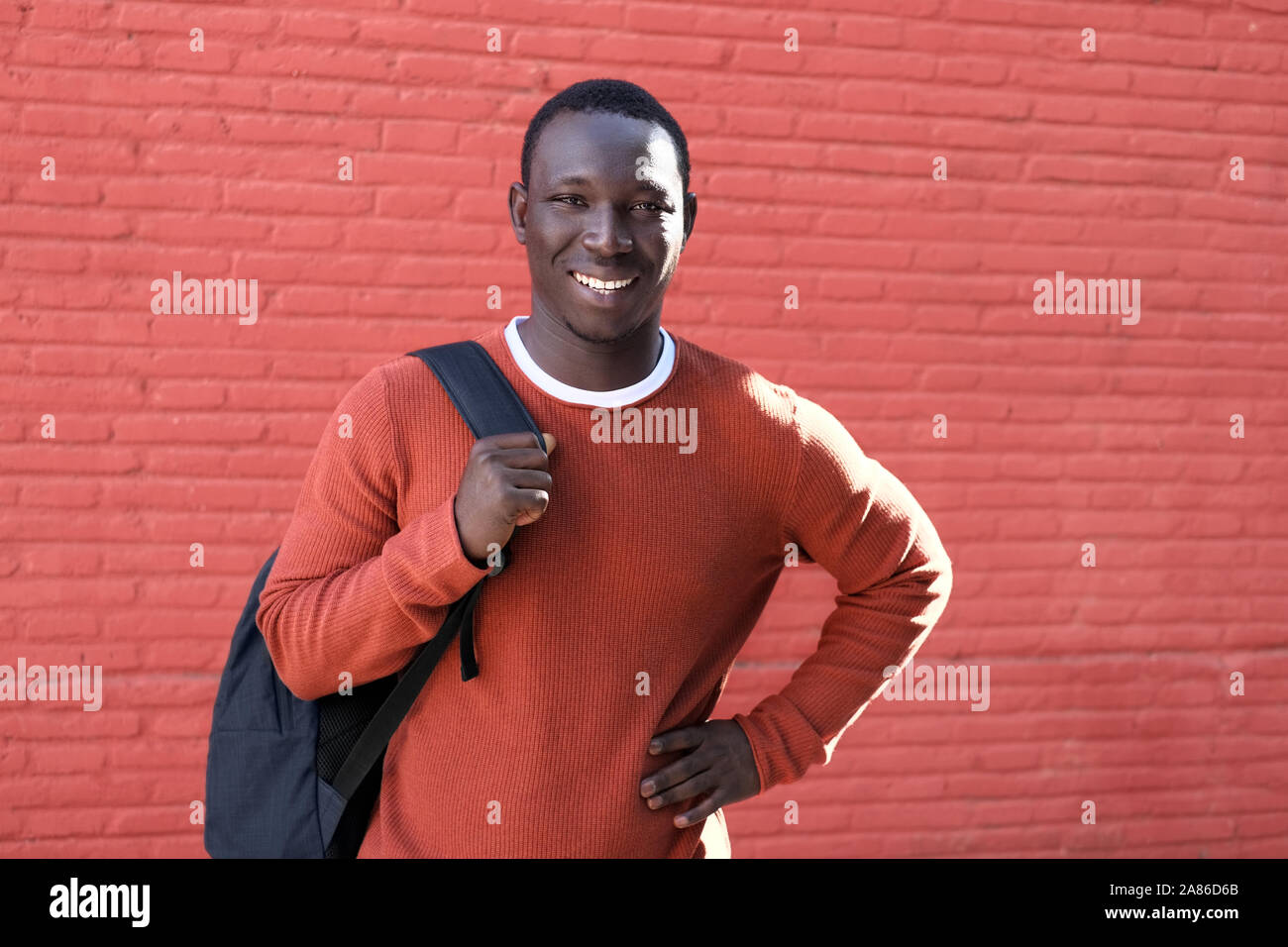 Portrait der Afrikanischen jungen Mann mit Kamera und lächelnd, mit Rucksack für die Schule. Kopieren Sie Speicherplatz auf rote Wand im Hintergrund. Stockfoto