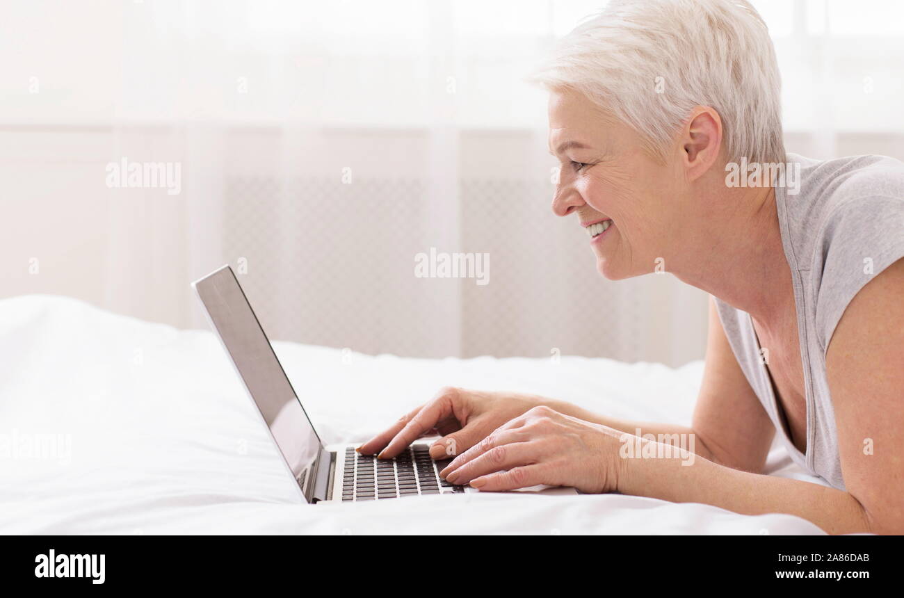 Freundliche ältere Dame Tippen auf Laptop, im Bett liegend Stockfoto
