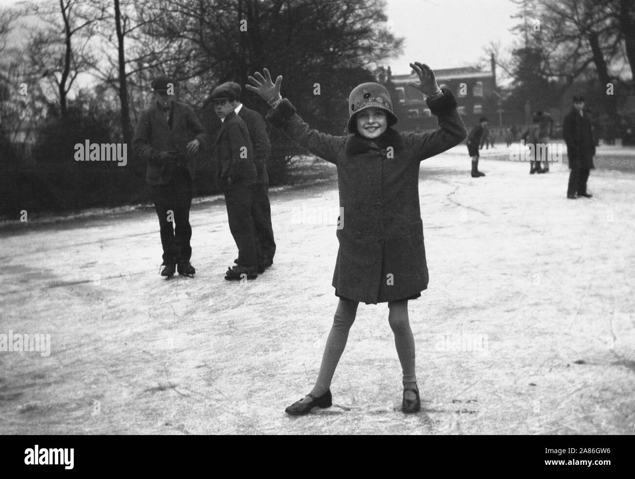 1930er Jahre, historisch, Kinder auf einer Eisfläche draußen, einige Jungs auf Schlittschuhen stehen zusammen und ein junges Mädchen in normalen Schuhen, Strumpfhosen, Mantel und Hut, Arme hoch, England, UK. Stockfoto