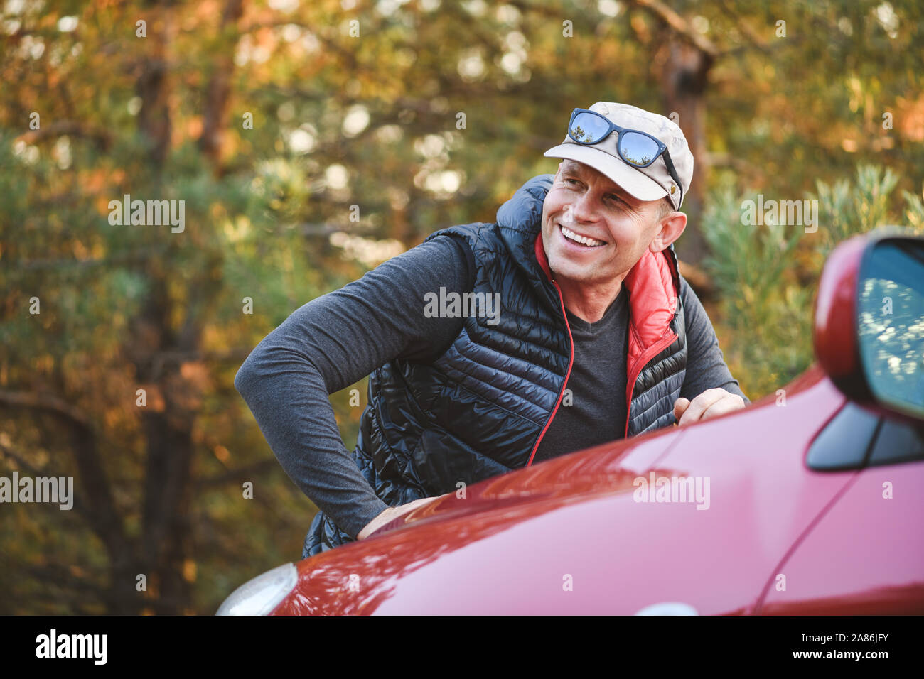 Lächelnd Mann lehnt sich auf der Motorhaube von einem roten Auto. SUV im Wald. Stockfoto