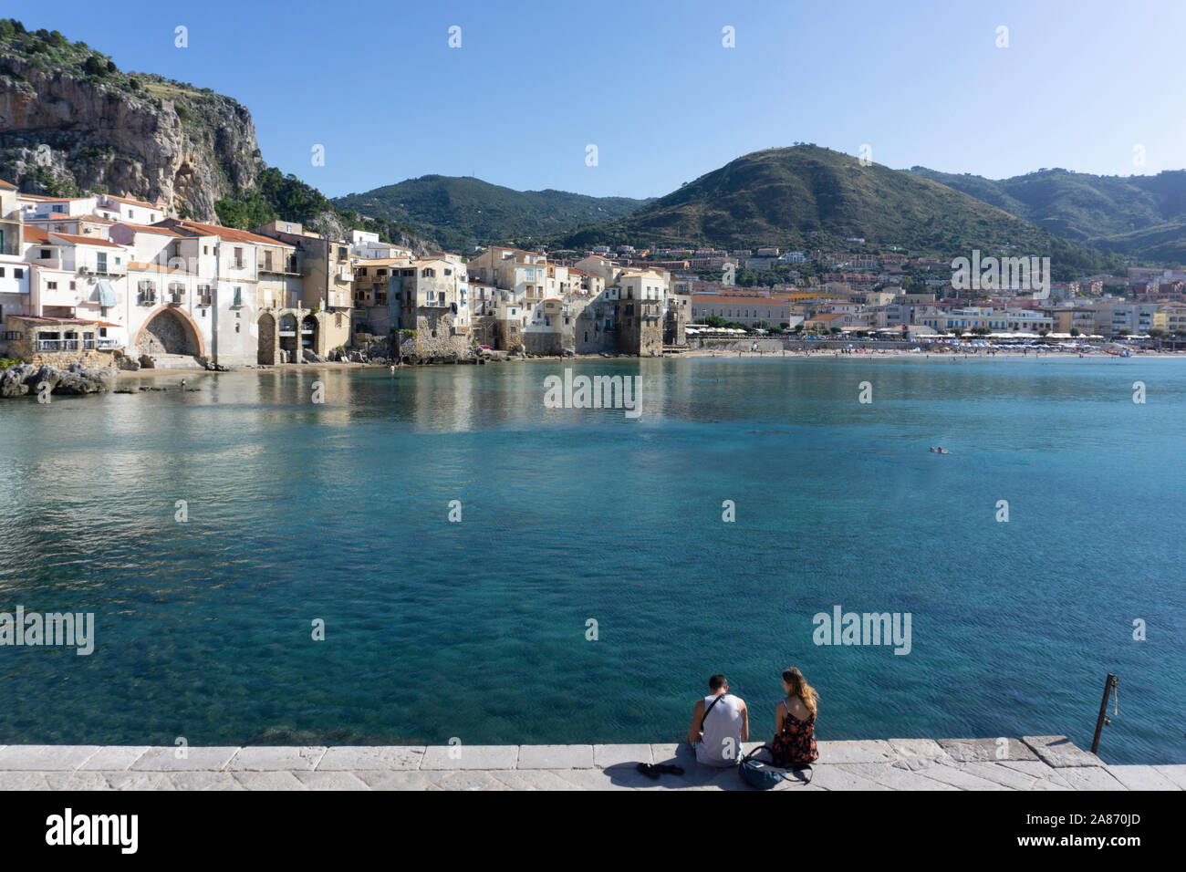 Ein paar sitzen am Pier, gegenüber der Altstadt von Cefalú, Sizilien, Italien, mit dem Felsen von La Rocca in der Stadt auftauchen. Stockfoto