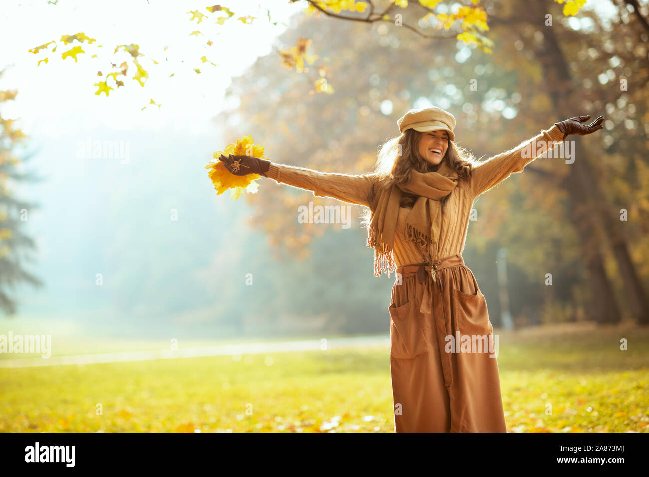 Hallo Herbst. lächelnd stilvolle Frau, Pullover, Rock, Mütze, Handschuhe und Schal mit gelben Blättern mit erhobenen Armen Jubel außerhalb im Herbst Park. Stockfoto
