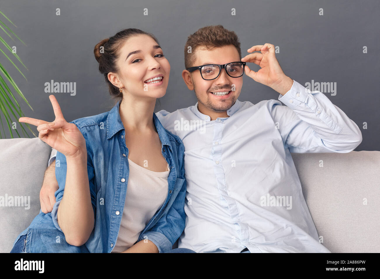 Relationnship Konzept. Junges Paar Mann in Gläsern und Frau, Frieden Geste sitzt auf einem Sofa studio isoliert auf Grau lächelt glücklich Stockfoto