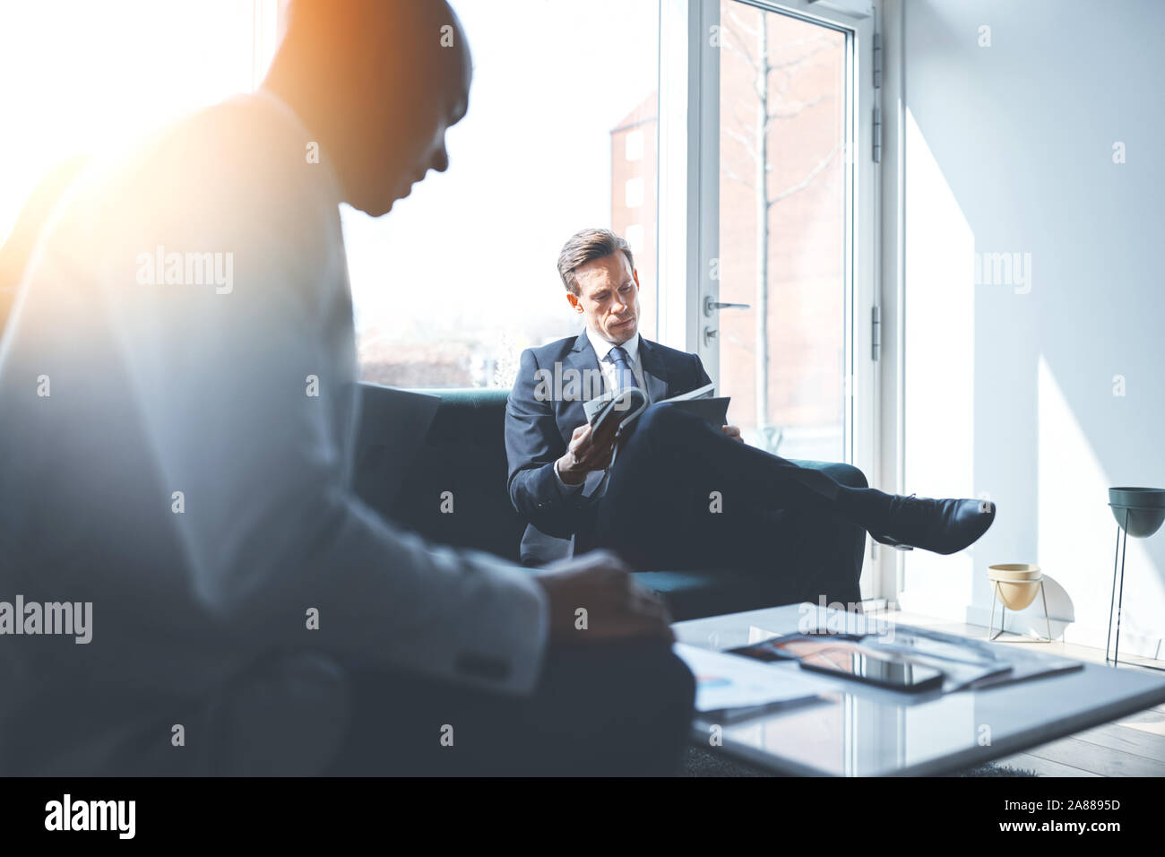 Geschäftsmann Lesen einer Zeitschrift beim Sitzen auf einem Sofa mit einem Kollegen in einem hellen modernen Büro Stockfoto