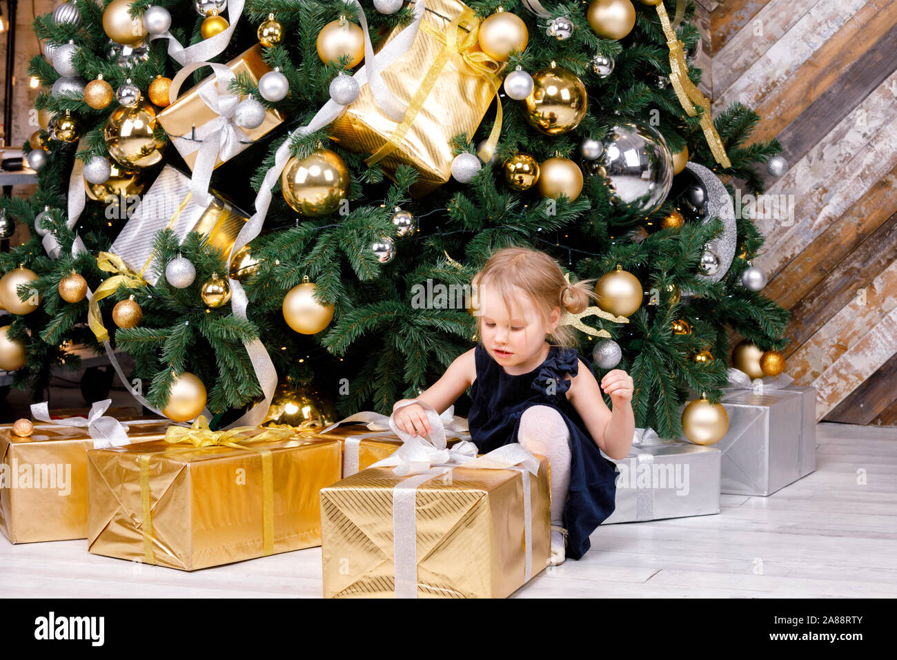 Portrait von elementare Alter Mädchen mit blauen Kleid neben Weihnachtsbaum auspacken Big verpackt Geschenkkarton neugierig auf ein Geschenk an Weihnachten sitzen Stockfoto