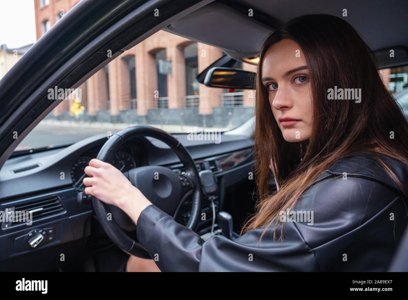 Eine junge Frau sitzt in ihrem Auto auf dem Parkplatz. Auf junge Mädchen leder Frauen schwarze Farbe. Konzept der Mädchen mit Charakter und gefährliches Fahren. Stockfoto