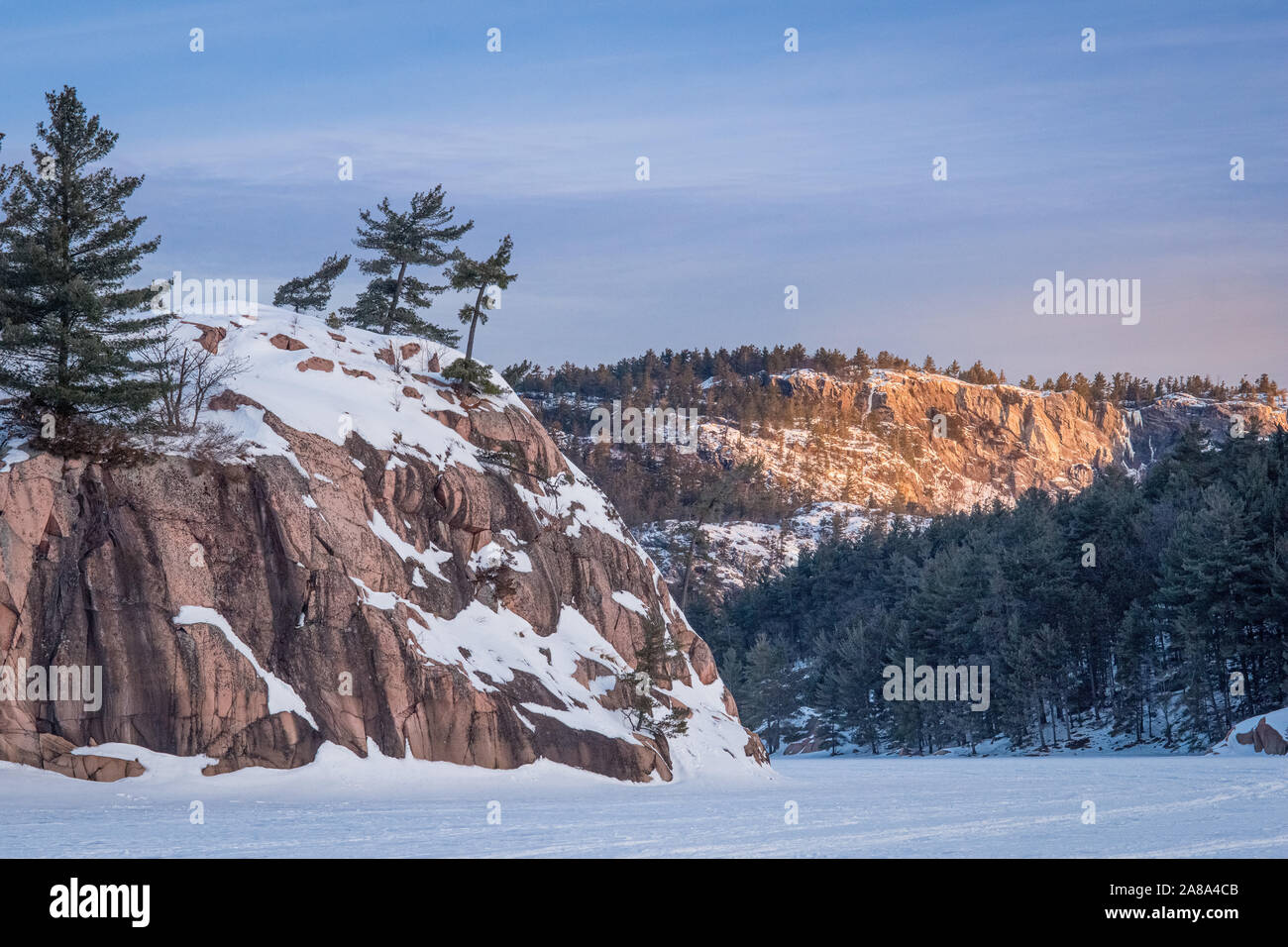 Dramatische orange dawn Sonnenlicht Streiks weit bergigen Ridge mit einem Wald und Felsvorsprung mit Bäumen im Vordergrund neben einem gefrorenen See Stockfoto