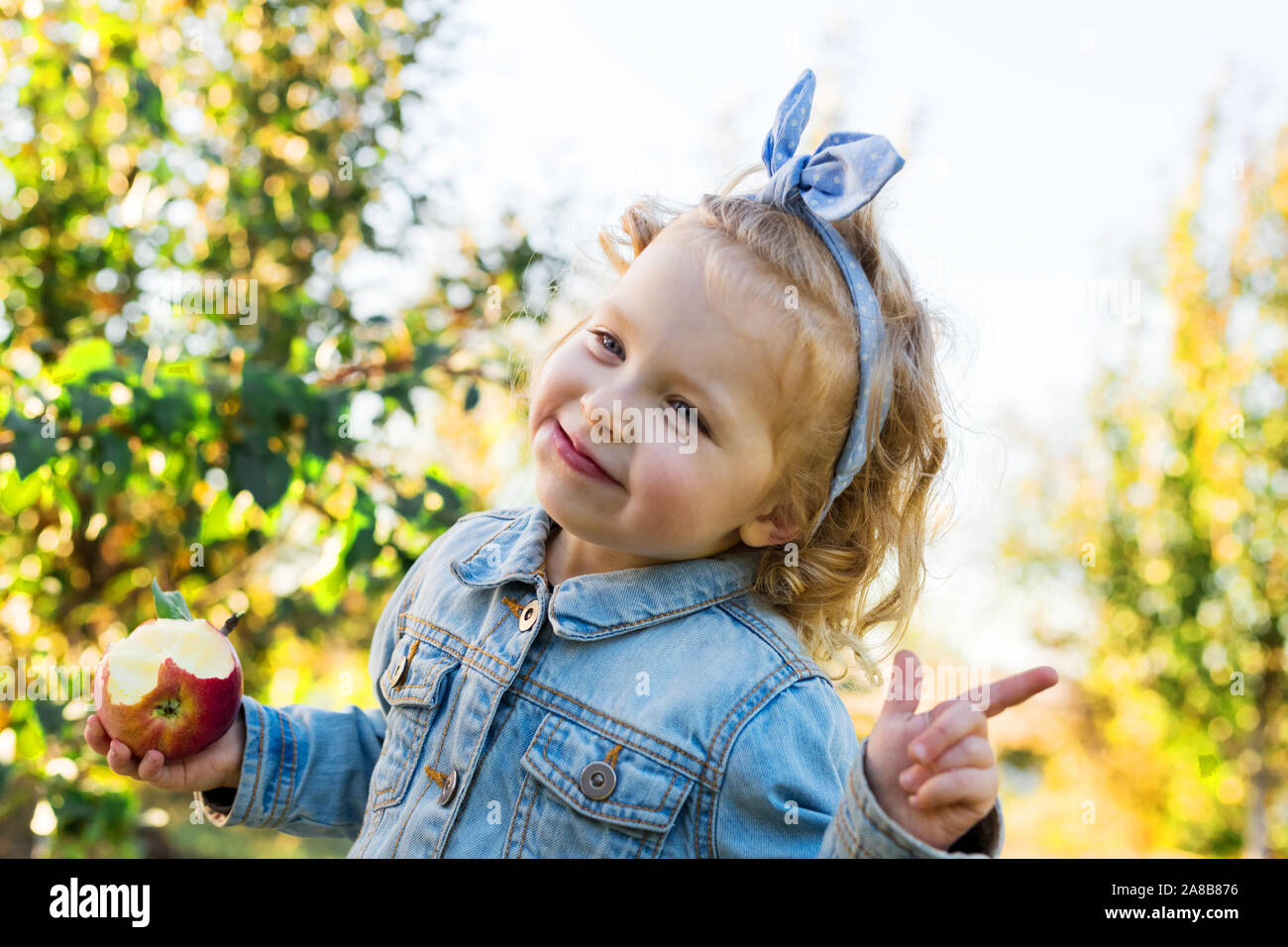 Süße kleine Mädchen essen Reif organische Red Apple im Apple Orchard im Herbst. Fair lockigen Haaren europäische Mädchen Kind in einer Jeans Anzug auf einem Bauernhof Stockfoto