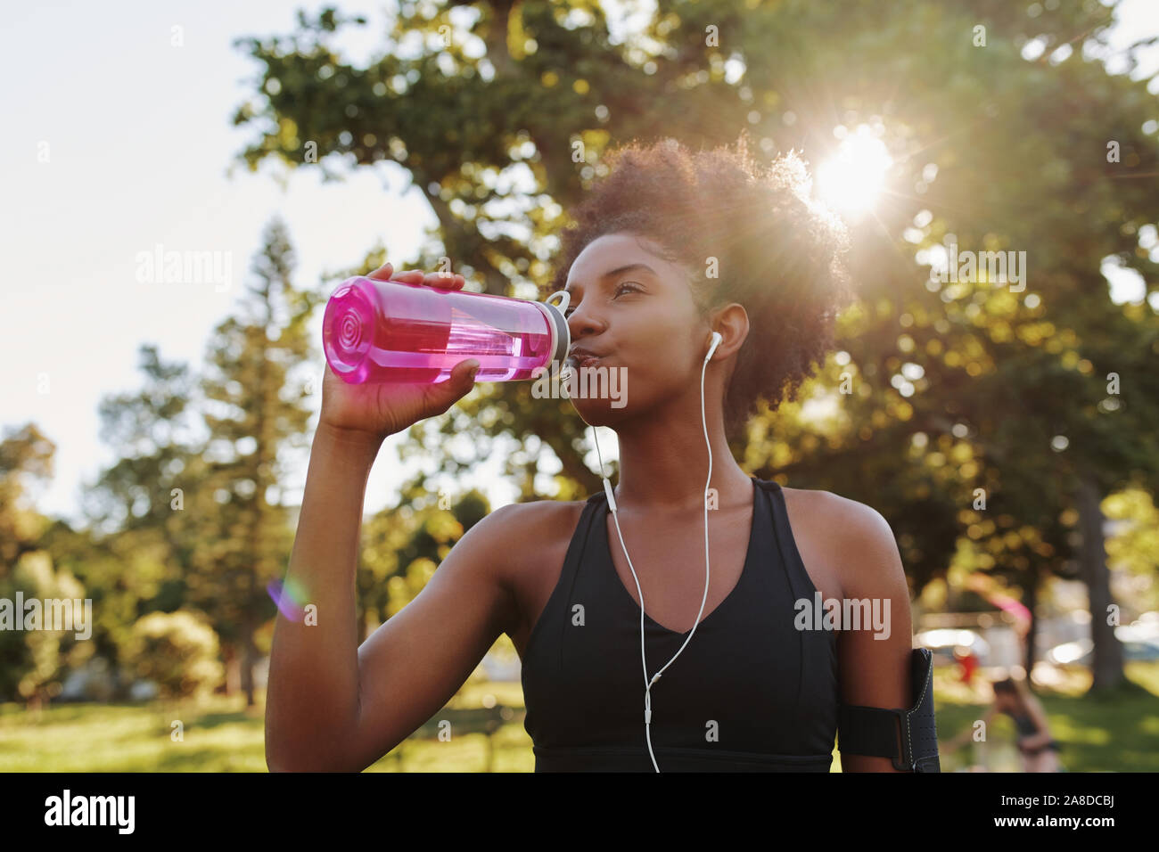 Fitness Sportler junge afrikanische amerikanische Frau Hören von Musik über Kopfhörer Trinkwasser in eine wiederverwendbare Trinkflasche nach Ausübung auf Stockfoto