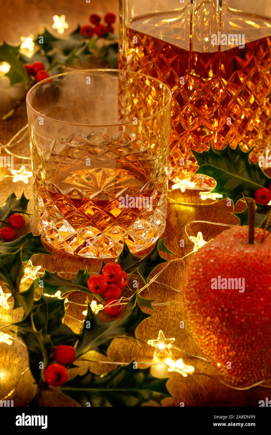 Kristall Glas Whisky und ein Whisky Karaffe mit Weihnachtsbeleuchtung Spielereien und Stechpalme Beeren, auf einem Holztisch Stockfoto