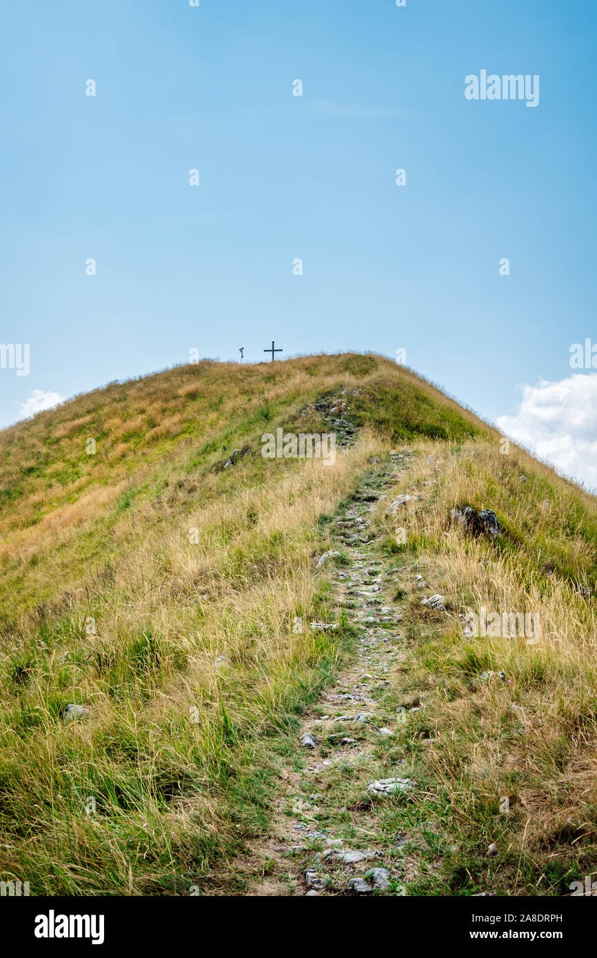 Wanderweg, auf einem Hügel mit Gras zu einem Berg mit einem Kreuz auf, gegen einen klaren blauen Himmel. Monte Boglia Peak bei Sommer, Schweizer Alpen. Stockfoto