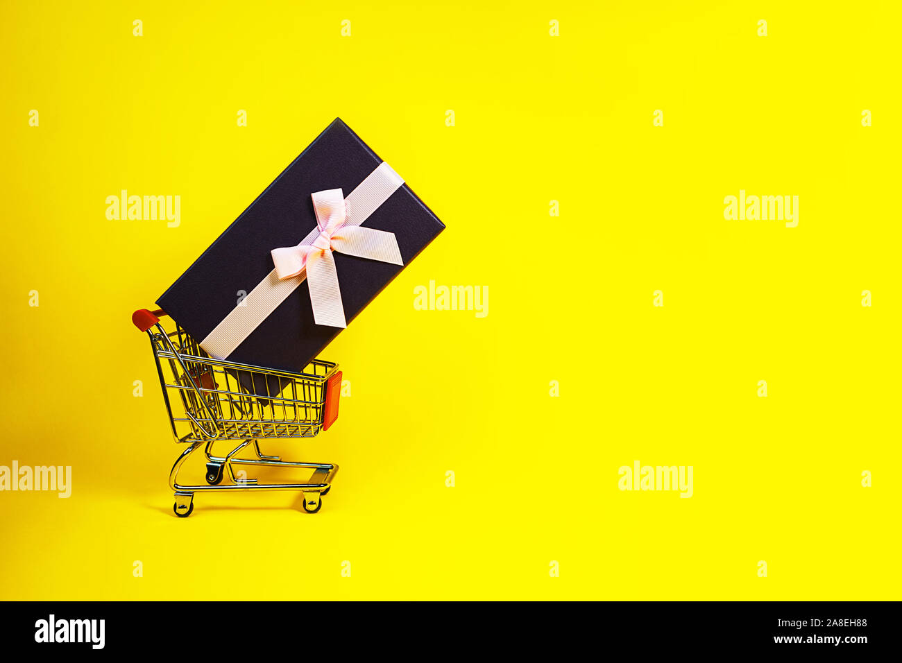 Urlaub Geschenk, Schwarzer Freitag Konzept mit Geschenkbox und rosa Schleife in einem Metall Einkaufswagen, Korb auf gelben Hintergrund. Shopping. Outfit. Flach, Ansicht von oben, Platz für Text. Stockfoto