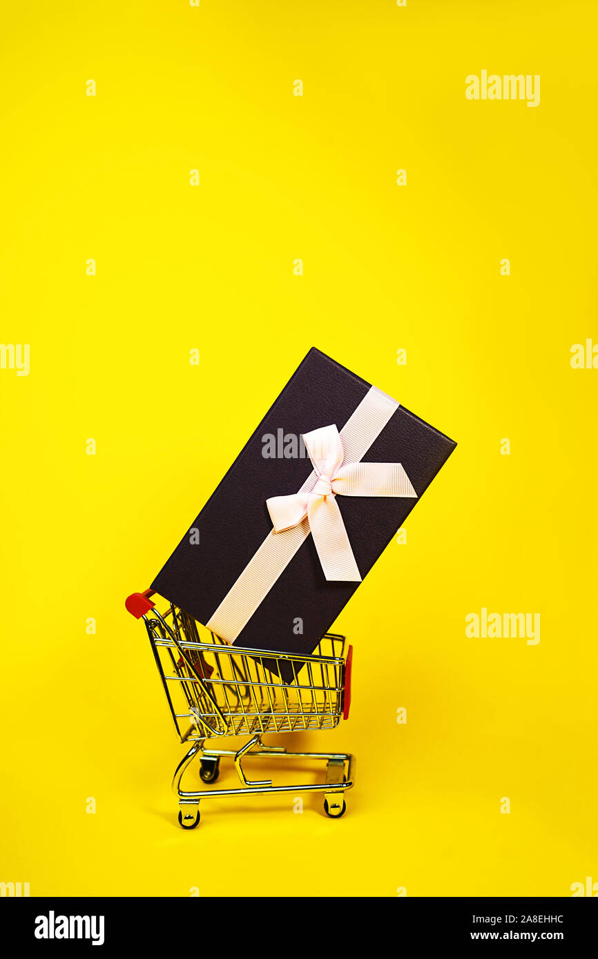 Urlaub Geschenk, Schwarzer Freitag Konzept mit Geschenkbox und rosa Schleife in einem Metall Einkaufswagen, Korb auf gelben Hintergrund. Shopping. Outfit. Flach, Ansicht von oben, Platz für Text. Stockfoto