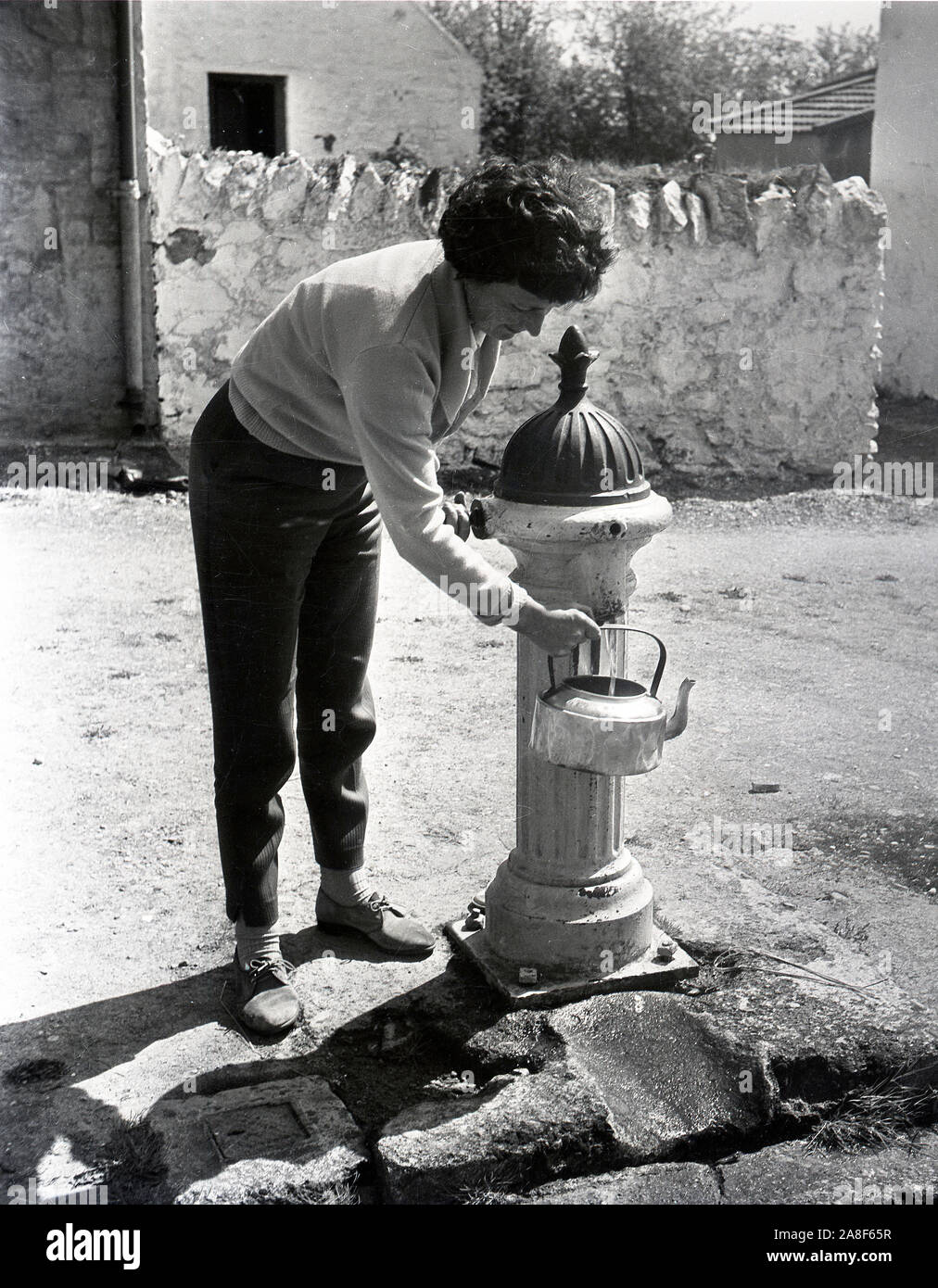 Frau Füllung Wasserkocher vom Dorf Gusseisen stehen Pumpe Großbritannien 1958 Stockfoto