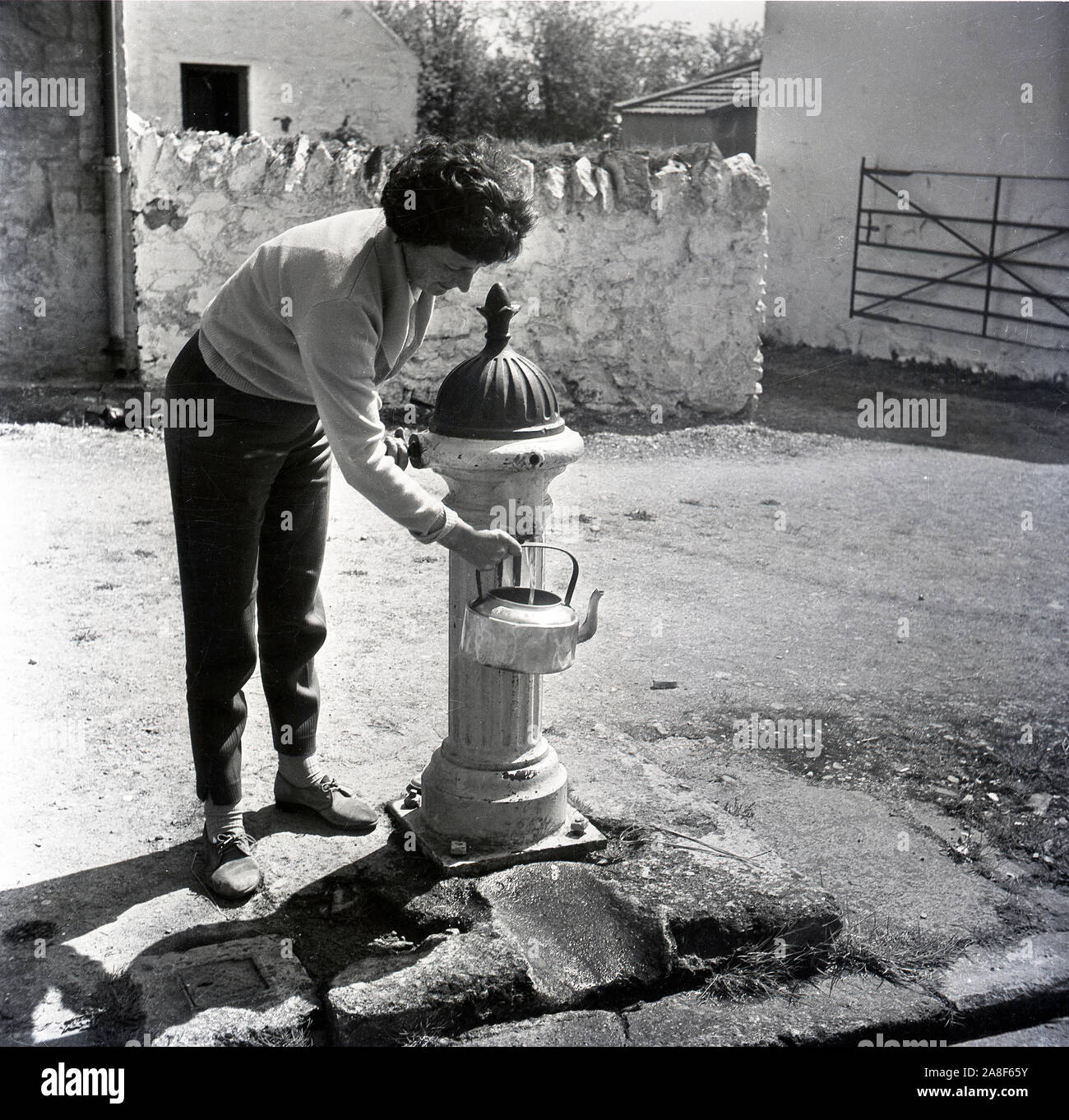 Frau Füllung Wasserkocher vom Dorf Gusseisen stehen Pumpe Großbritannien 1958 Stockfoto