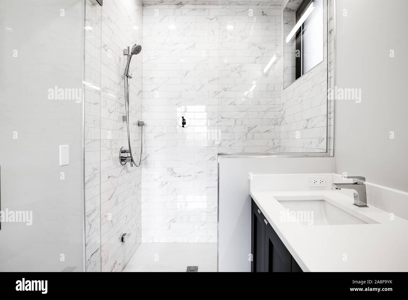 Ein luxuriöses Badezimmer mit weißem Granit auf einer dunklen Eitelkeit und weißem Marmor gefliest mit Glaswänden und Duschtür. Stockfoto