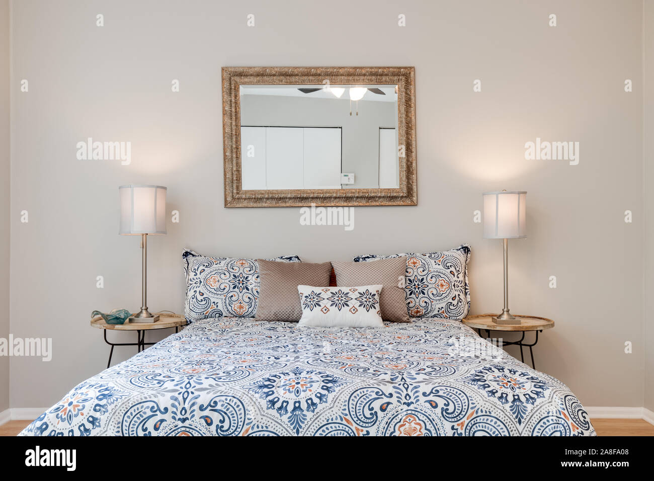 Ein Bett im Schlafzimmer zu einer Residenz zentriert mit zwei Lampen auf dem Nachttisch und ein Spiegel hängt über dem Bett. Stockfoto