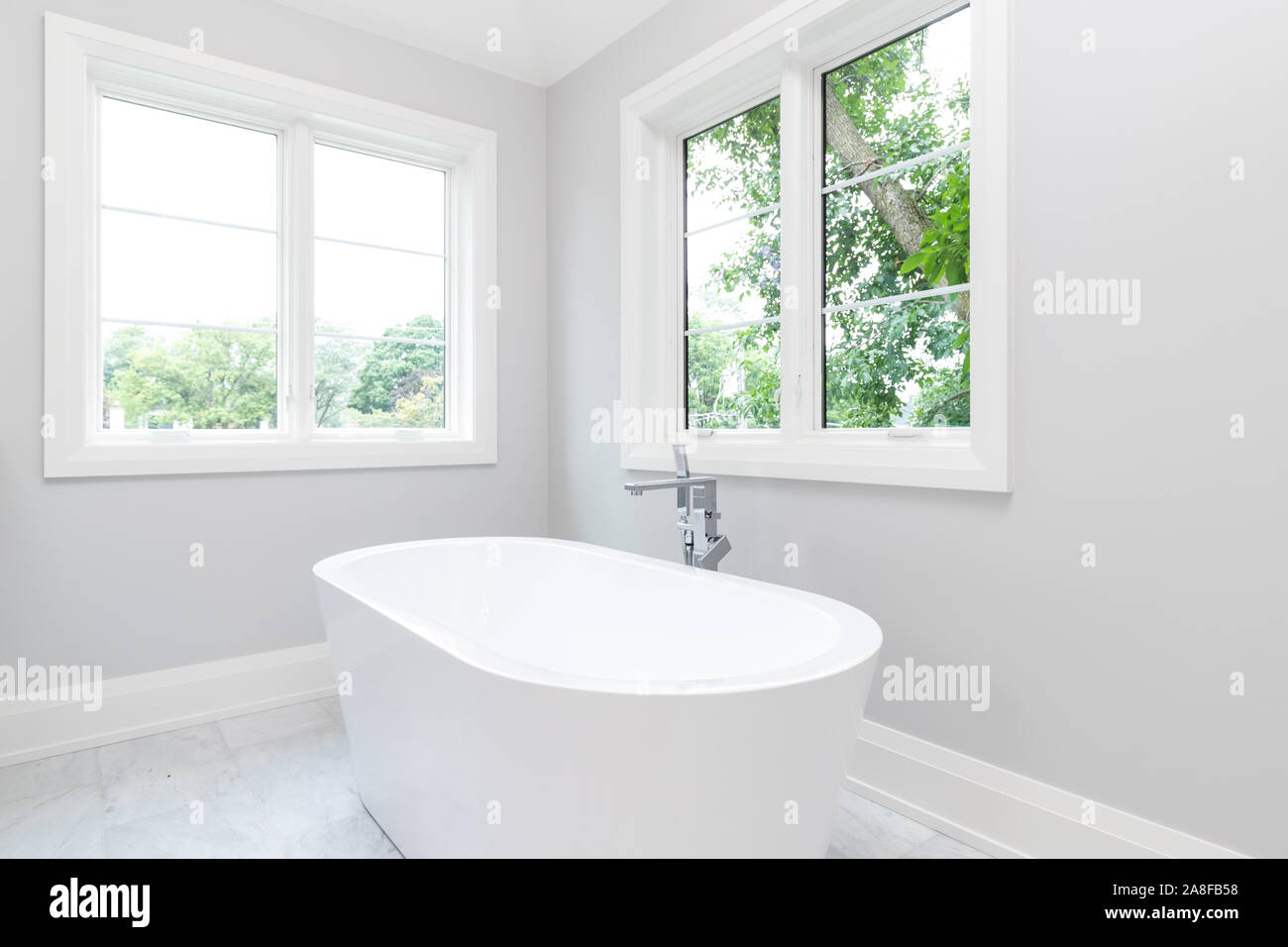 Eine schöne, weiße standalone Badewanne mit Chrom Hardware und schönen grünen Bäumen werden aus den Fenstern zu sehen. Stockfoto