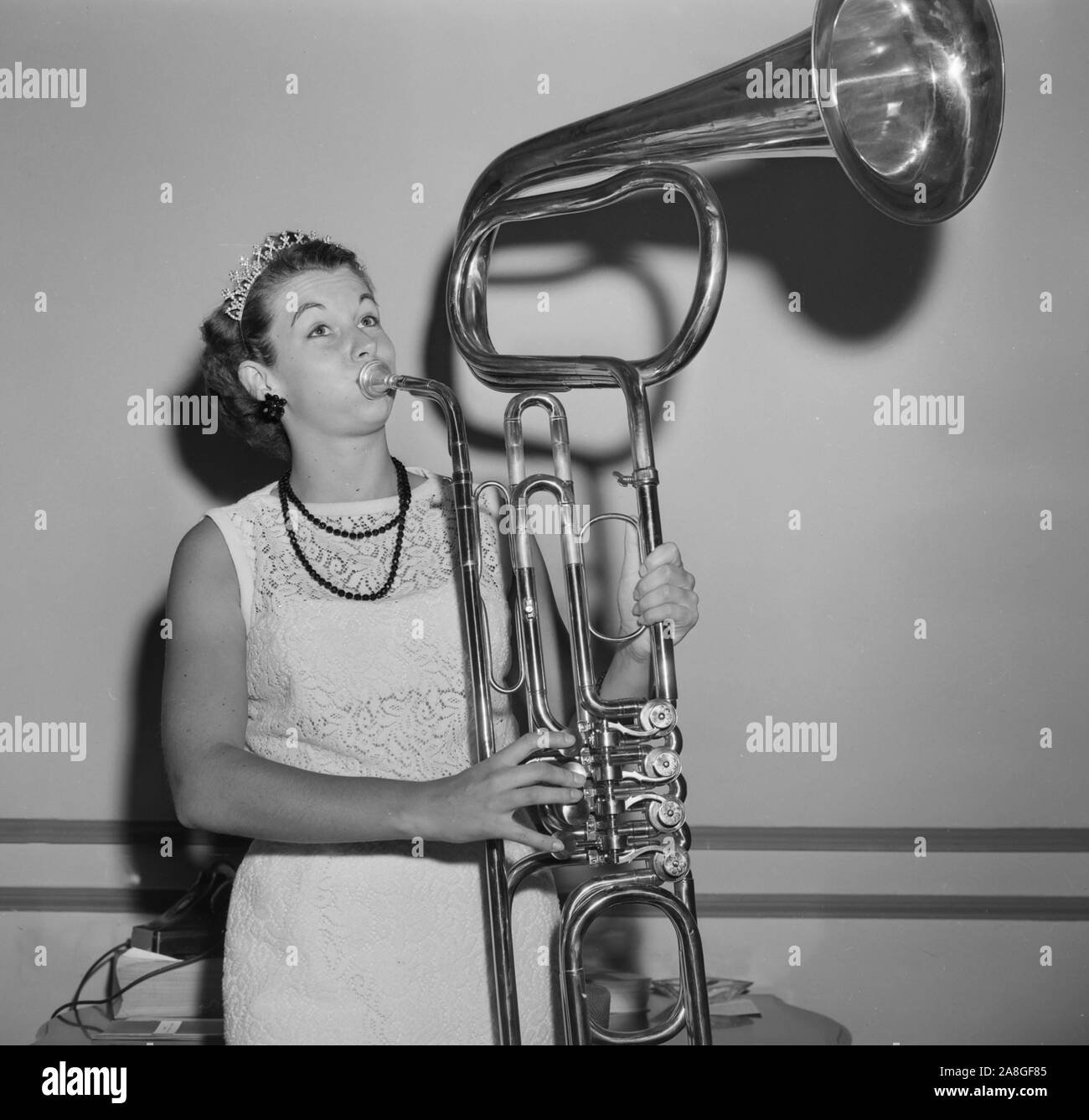 Eine junge Frau, vielleicht ein schönheitswettbewerb Kandidat, bläst einem ungewöhnlich konstruierte Horn aus Stücken von anderen Band Instrumenten gemacht, Ca. 1960. Stockfoto