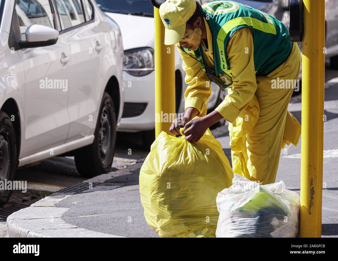 Afrikanische garbage collector bindet einen Knoten in einer vollen gelben Kunststoff Müllsack auf eine urbane Stadt Straße Ecke in Kapstadt, Südafrika Stockfoto