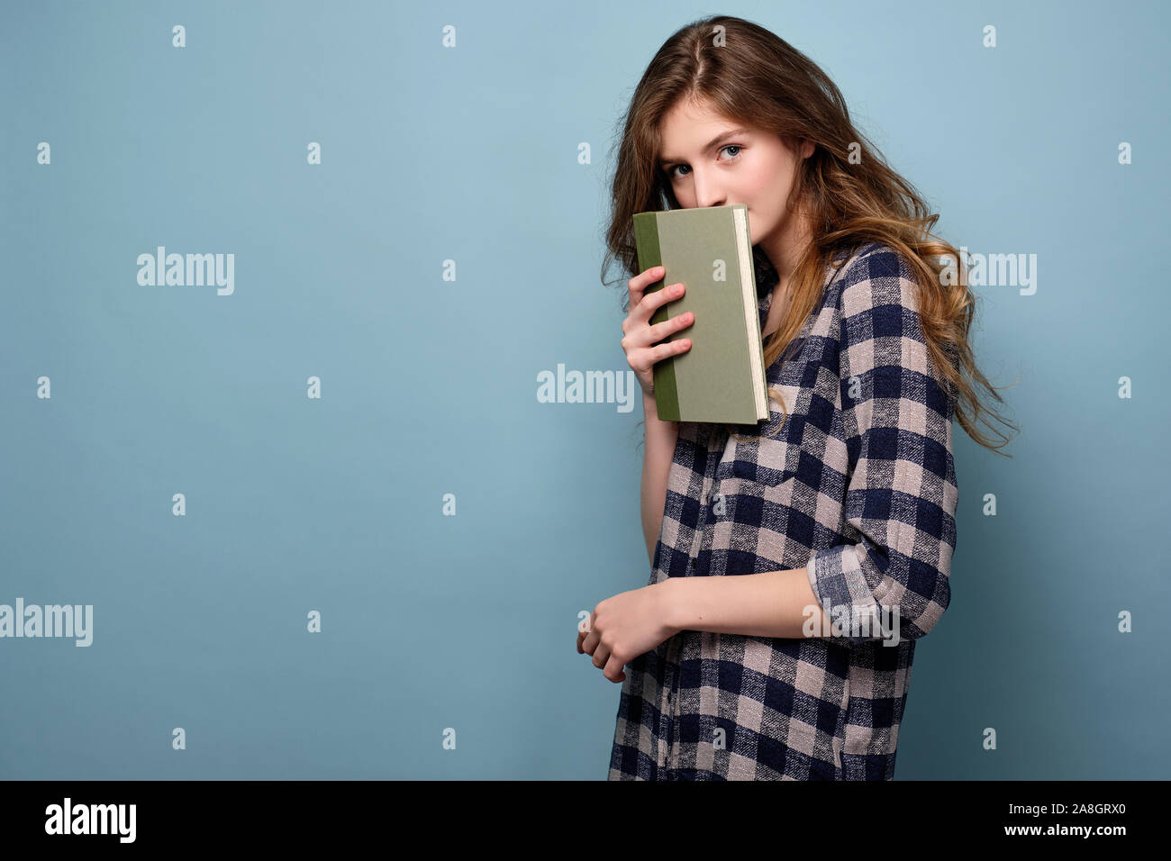 Ein Mädchen in einem Plaid Shirt steht in einer halben Umdrehung auf einem blauen Hintergrund und sieht in den Rahmen, hinter einem Buch versteckt. Horizontale Foto Stockfoto