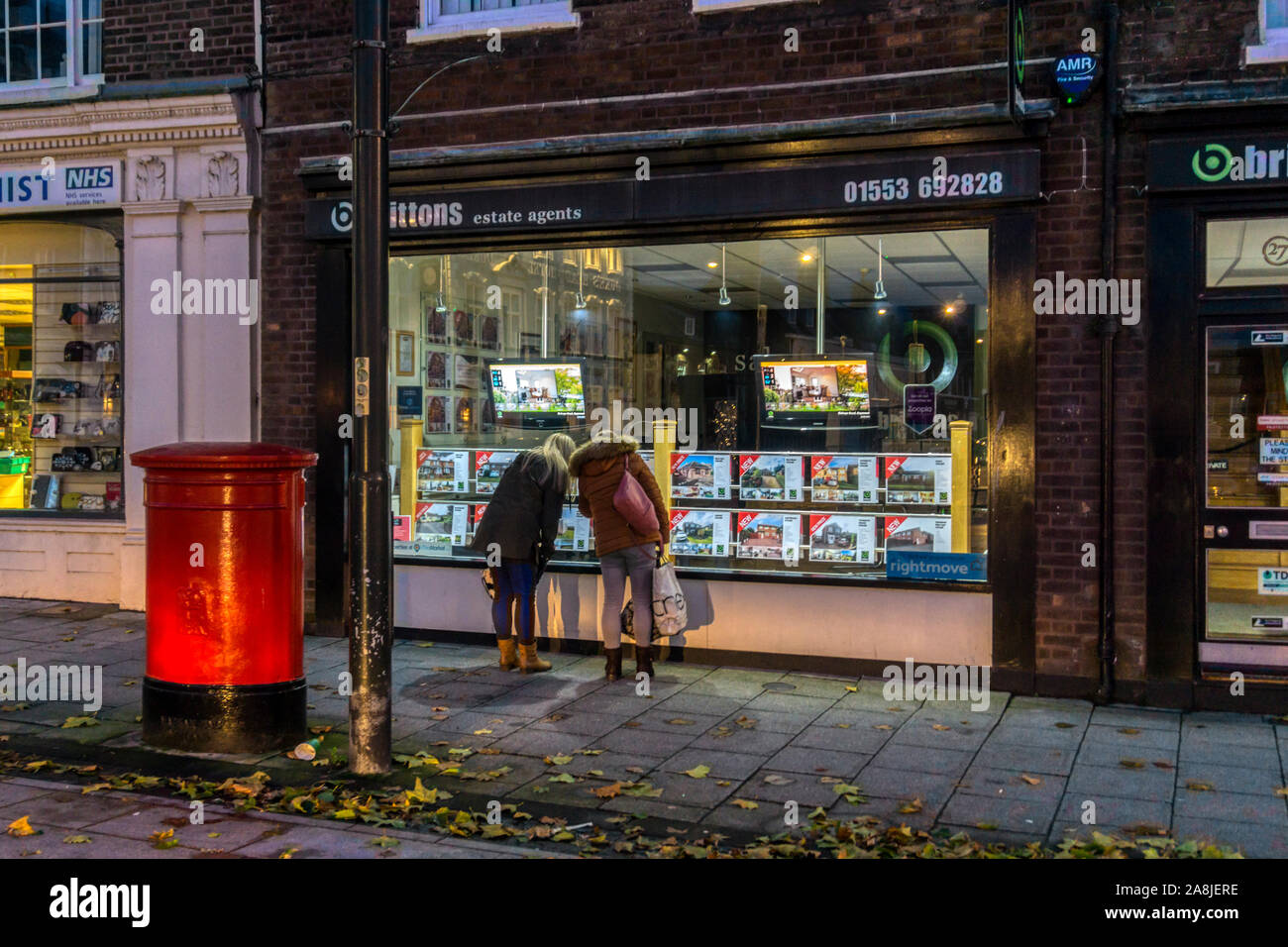 Zwei Frauen auf der Suche nach Details in einem beleuchteten Fenster der Immobilienmakler in der Nacht, auf Kings Lynn Dienstag Markt. Stockfoto