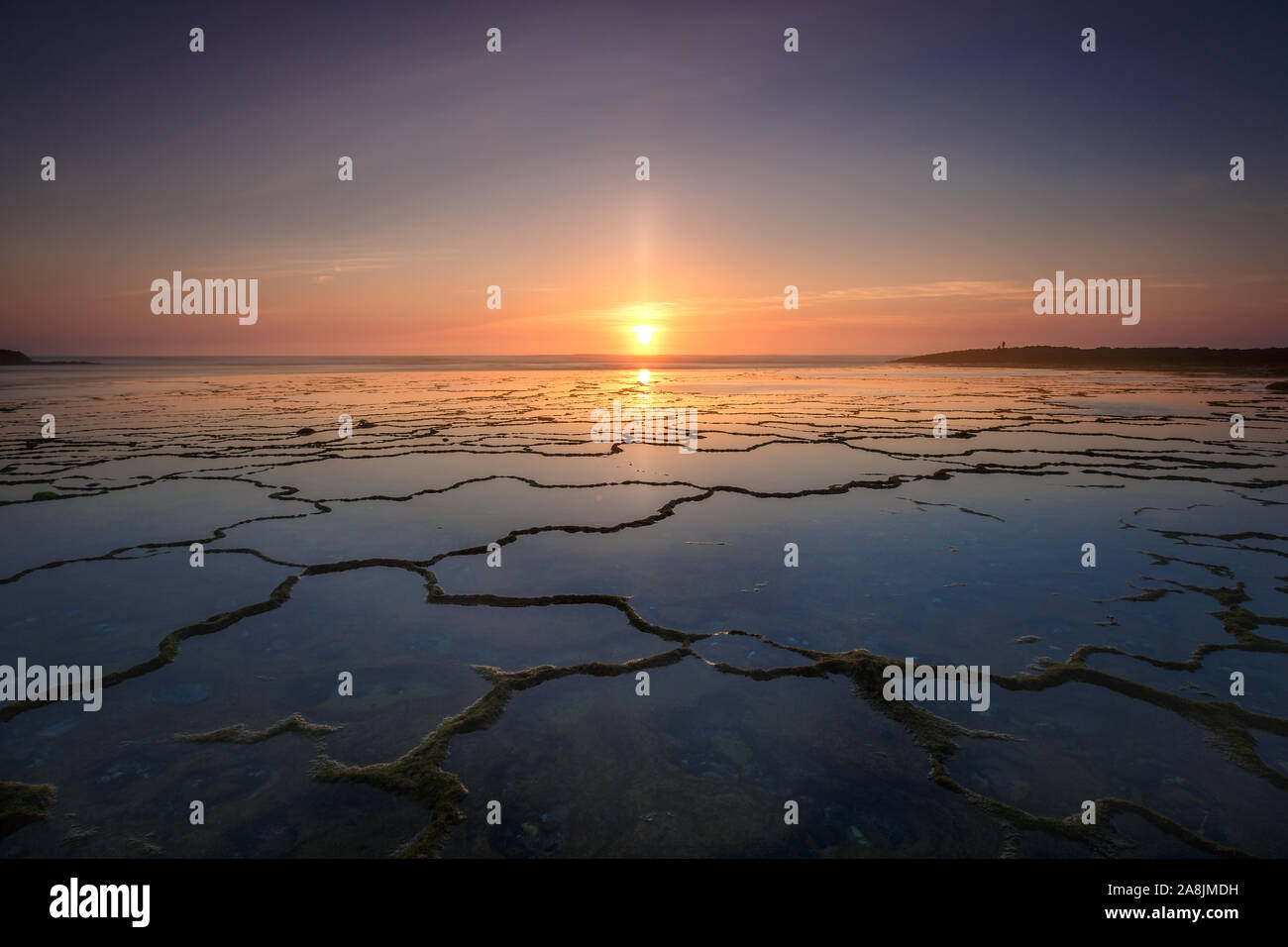 Sonnenuntergang mit Solar pilar Phänomene im Praia do Farol Strand, im Alentejo, Portugal. Erste Ebene der Felsformation mit Net Shape. Wasser spiegelt sich der Himmel Stockfoto