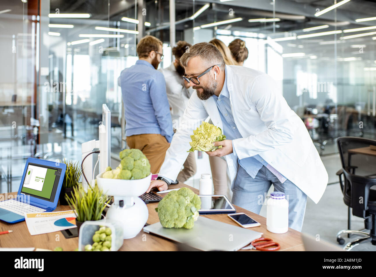 Leitende Ernährungsberaterin arbeiten auf einer Diät Plan am Arbeitsplatz mit Computer und frische Lebensmittel im Büro mit jungen Kolleginnen und Kollegen im Hintergrund Stockfoto