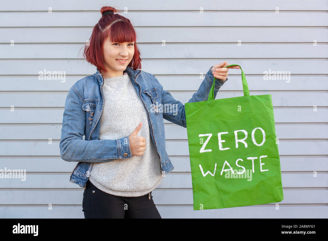 Mädchen zeigt Daumen hoch halten eine Tasche mit der Aufschrift null Abfall. Das Mädchen trägt eine Jeans Jacke und Hose. Grauer Hintergrund. Stockfoto