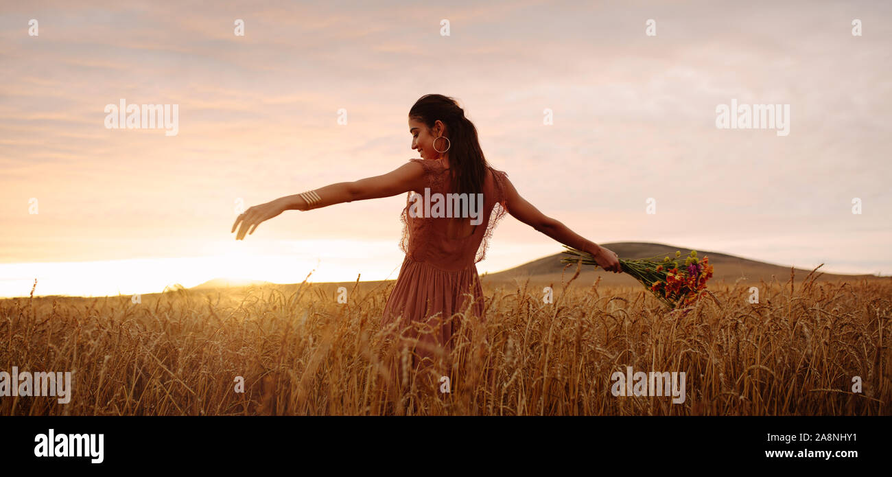 Junge Frau im Weizenfeld holding Blumenstrauß und bei Sonnenuntergang zu genießen. Weibchen mit ihren ausgestreckten Armen stehend in der ursprünglich eingereichten Fassung mit Stockfoto