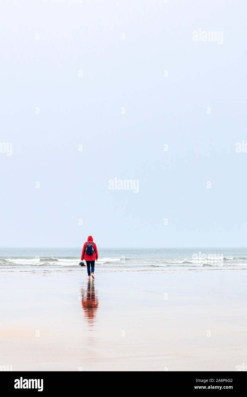 Eine einsame Figur in einem roten Anorak zu Fuß in Richtung Meer an einem regnerischen und windigen Strand, für ein Buch geeignet, Westward Ho!, Devon, Großbritannien Stockfoto