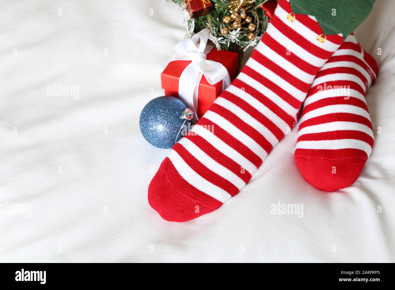 Weihnachtsfest zu Hause, überquerte weibliche Füße in rot-weiß gestreifte Socken und Neues Jahr Baum auf einem Bett. Frau in Santa Kleidung und festliche Dekoration Stockfoto