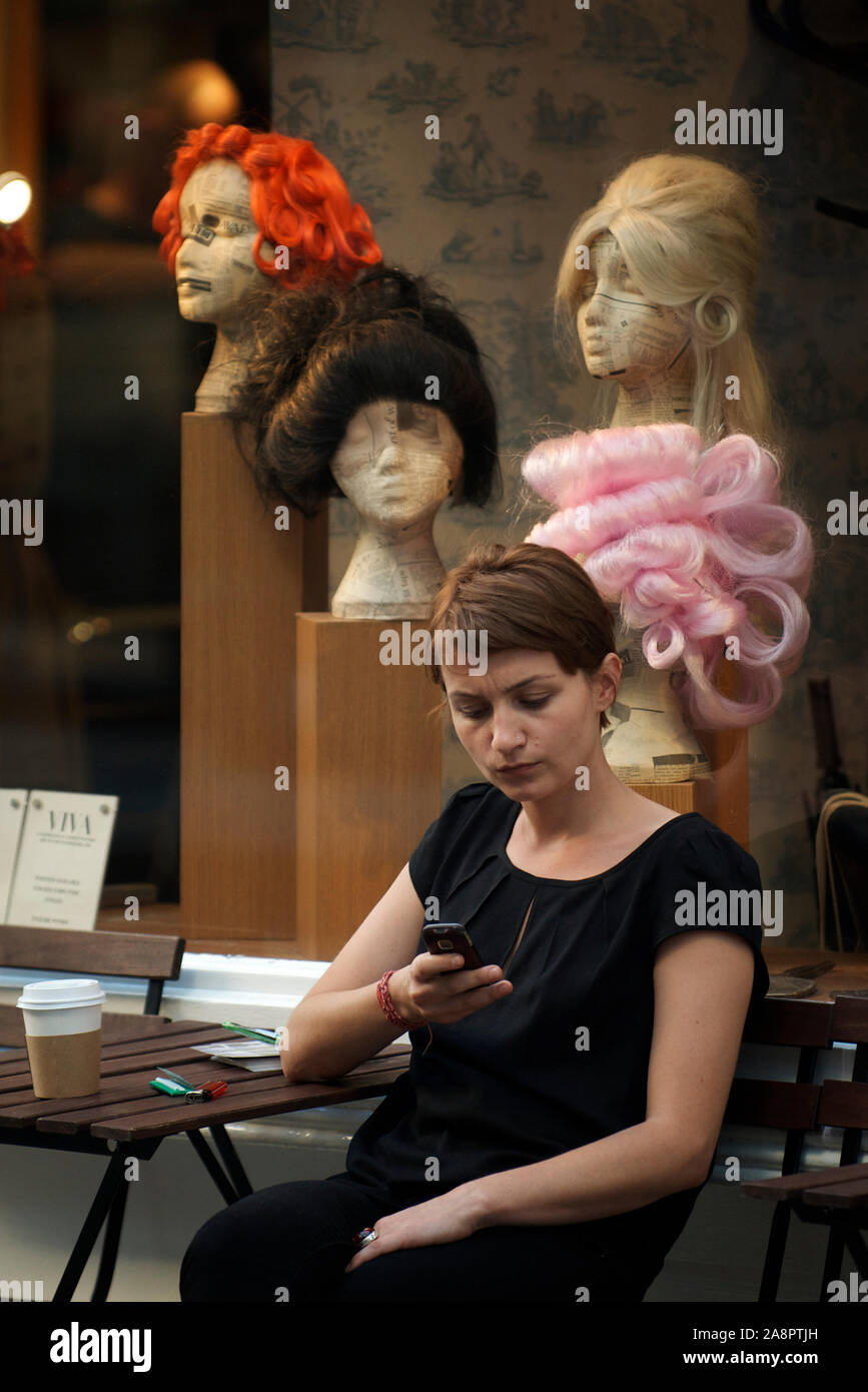 London - 30. SEPTEMBER 2011: Eine junge Frau sitzt draußen mit ihrem Handy vor einem Schaufenster mit bunten Perücken in Soho. Stockfoto