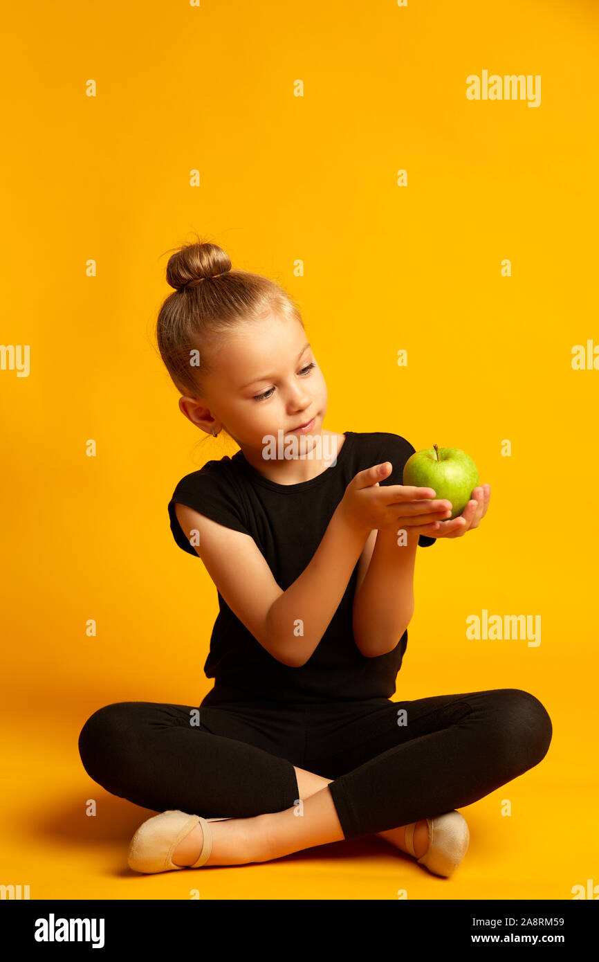 Voller Körper kleines Mädchen in leotard an grünen Apfel, während gegen gelben Hintergrund während der Pause in der Ausbildung sitzen Stockfoto