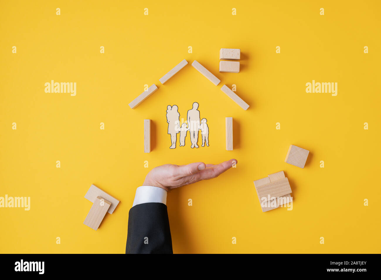 Geschäftsmann seine Hand unter einem Scherenschnitt Silhouette einer Familie in einem Haus aus Holz Blöcke und Stifte in einem konzeptionellen Bild. Über gelb zurück Stockfoto