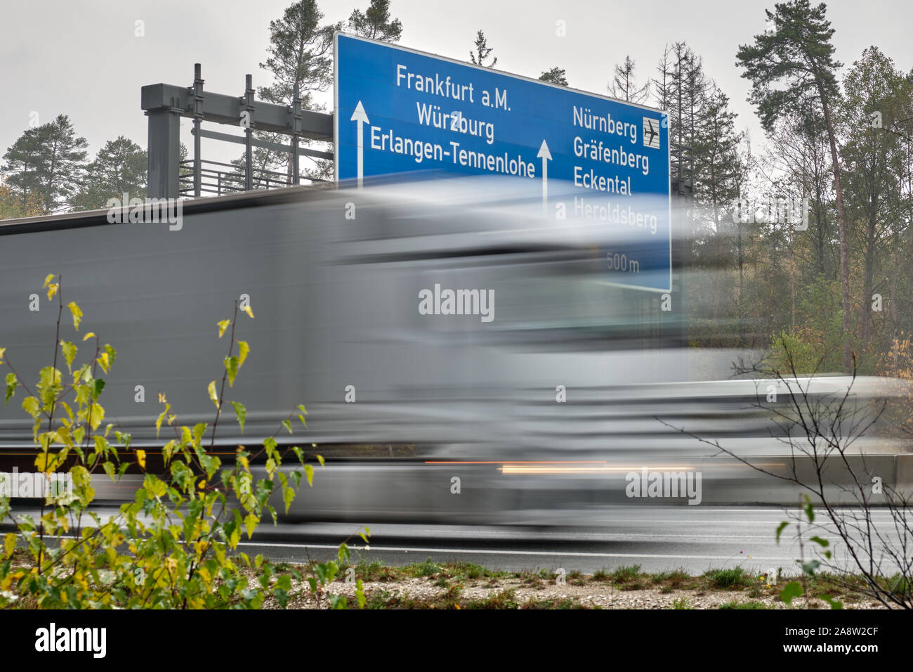 Lkw in verschwommene Bewegung entlang einer blauen Verkehrszeichen in Deutschland auf der Autobahn A3 zeigen den Weg nach Frankfurt am Main, Würzburg, Erlangen und N Stockfoto