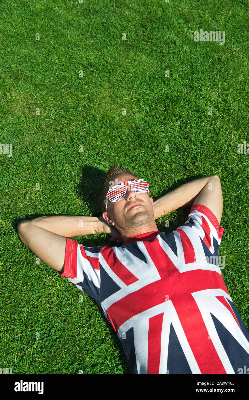 Patriotische junge Mann in Union Jack T-Shirt und Sonnenbrille Entspannen auf einem Flecken des Großen Britischen Gras Stockfoto