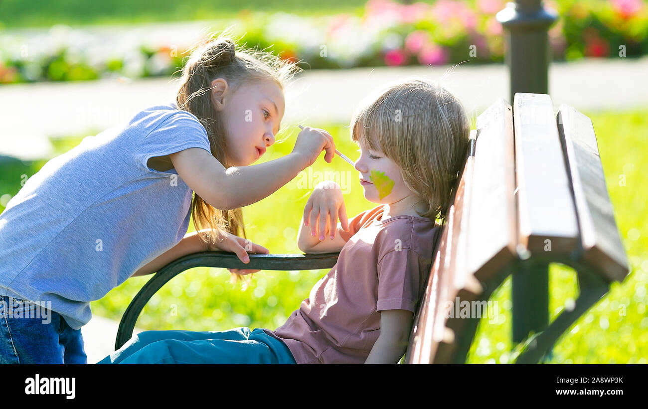 Ein kleines Mädchen ist die Anwendung aqua Make-up im Gesicht von einem 5 Jahre alten Jungen, der auf einer Bank sitzen ein Park ist. Stockfoto