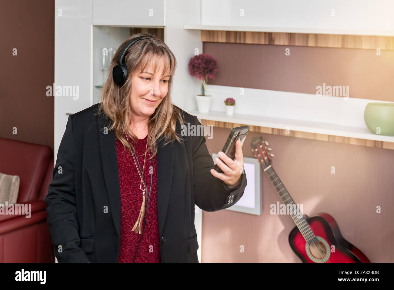 40-jährige Frau mit blonden Haaren liebt Musik über die Kopfhörer. Sie steuert die Musik mit dem Smartphone. Warme Farbe Konzept Herbst Winter. Stockfoto