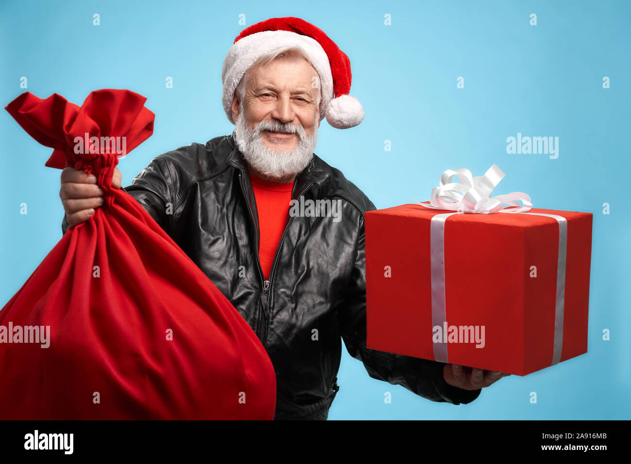 Fröhliche reifer Mann mit grauen Bart in Santa Hut und schwarzen Leder Jacke tragen viele Weihnachtsgeschenke in seinen Händen. Konzept der Winterurlaub und Feier. Stockfoto